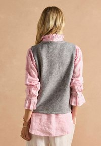 Grauer Wollwesten über einem rosa-weißen gestreiften Hemd mit Rüschensäumen. Weiche Texturen und geschichtetes Design sind sichtbar.