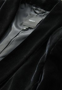 Blazer in velluto nero con foderatura interna in raso. Presenta un'etichetta con il marchio "NEXT" e un'etichetta cucita che indica "Fabbricato in Cina".