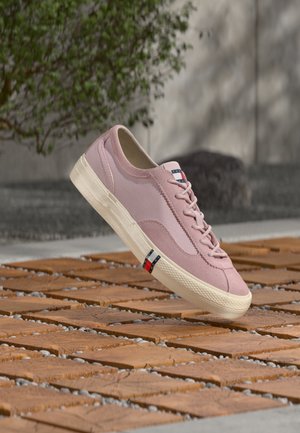 Lyserosa snøresko med hvit såle og detaljert rødt, hvitt og blått logo, plassert på et utendørs underlag av trebiter.