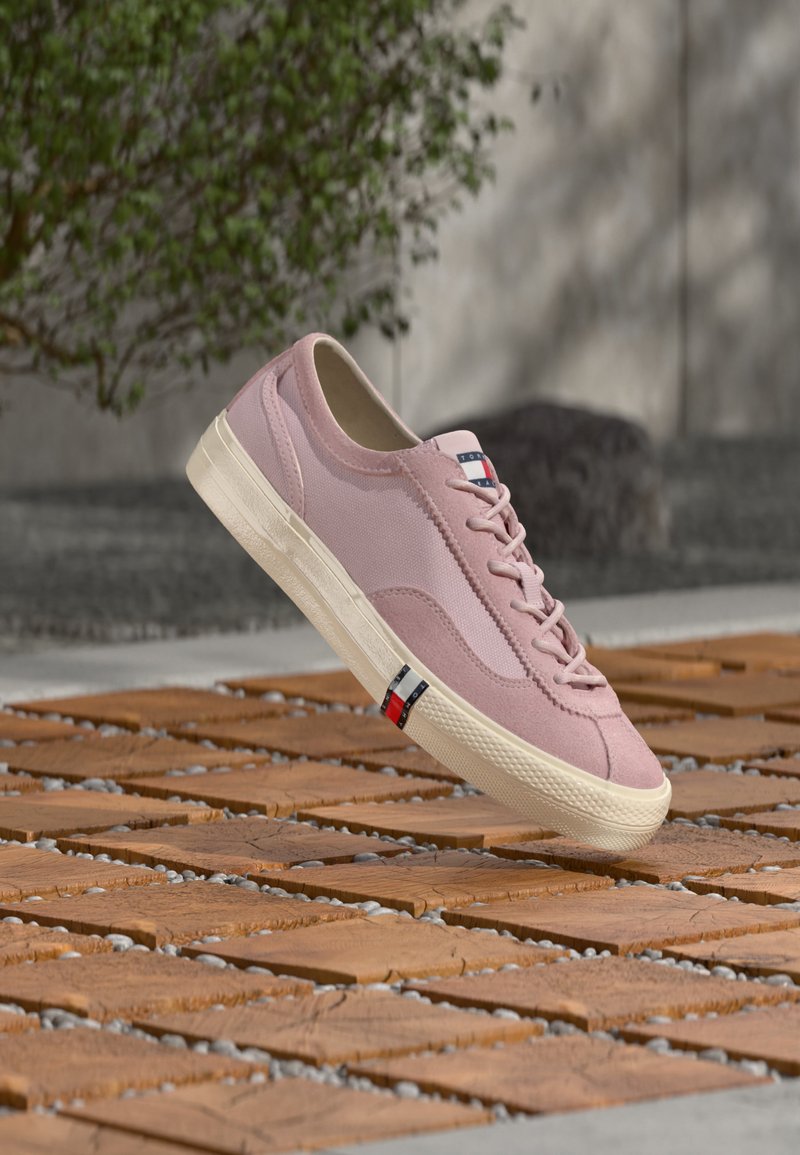 Ανοιχτό ροζ αθλητικό παπούτσι με κορδόνια, λευκή σόλα και λογότυπο σε κόκκινο, λευκό και μπλε, τοποθετημένο σε ξύλινη επιφάνεια πλακιδίων σε εξωτερικό χώρο.