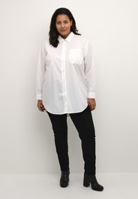 Camisa blanca de manga larga con botones y un único bolsillo en el pecho, combinada con pantalones negros ajustados y botines negros. Textura de tela suave.