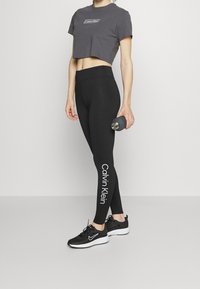 Zwarte leggings met het "Calvin Klein" logo aan de zijkant, gecombineerd met een cropped grijs t-shirt en zwarte sneakers. Houdt een waterfles vast.