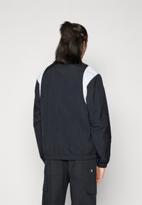 Lehká černá bunda s bílým akcentem na ramenou, žebrovanými manžetami a elastickým pasem. Texturovaná látka, minimalistický design.