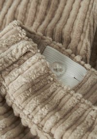 Tessuto beige strutturato con righe verticali, caratterizzato da una superficie morbida e pelosa. Include un'etichetta bianca con un dettaglio a bottone.