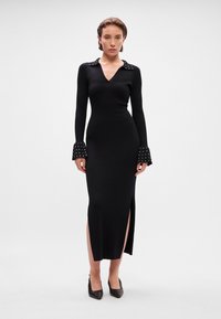 Vestido longo preto canelado com decote em V e colarinho de bolinhas. Possui mangas largas e uma fenda lateral para um detalhe adicional.