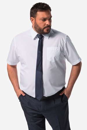 Man draagt een wit overhemd met korte mouwen, een donkerblauwe das en een donkere broek, staat met handen in de zakken en kijkt opzij tegen een effen achtergrond.