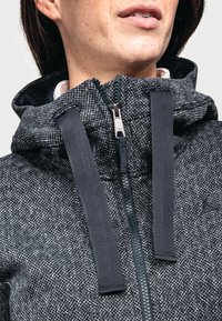 Chaqueta gris texturizada con capucha, que presenta un cierre frontal y correas oscuras en el cuello. Patrón de espiga notable en toda la prenda.