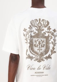 White cotton t-shirt featuring a large, distressed gold graphic of a crest and floral design on the back, with text below.