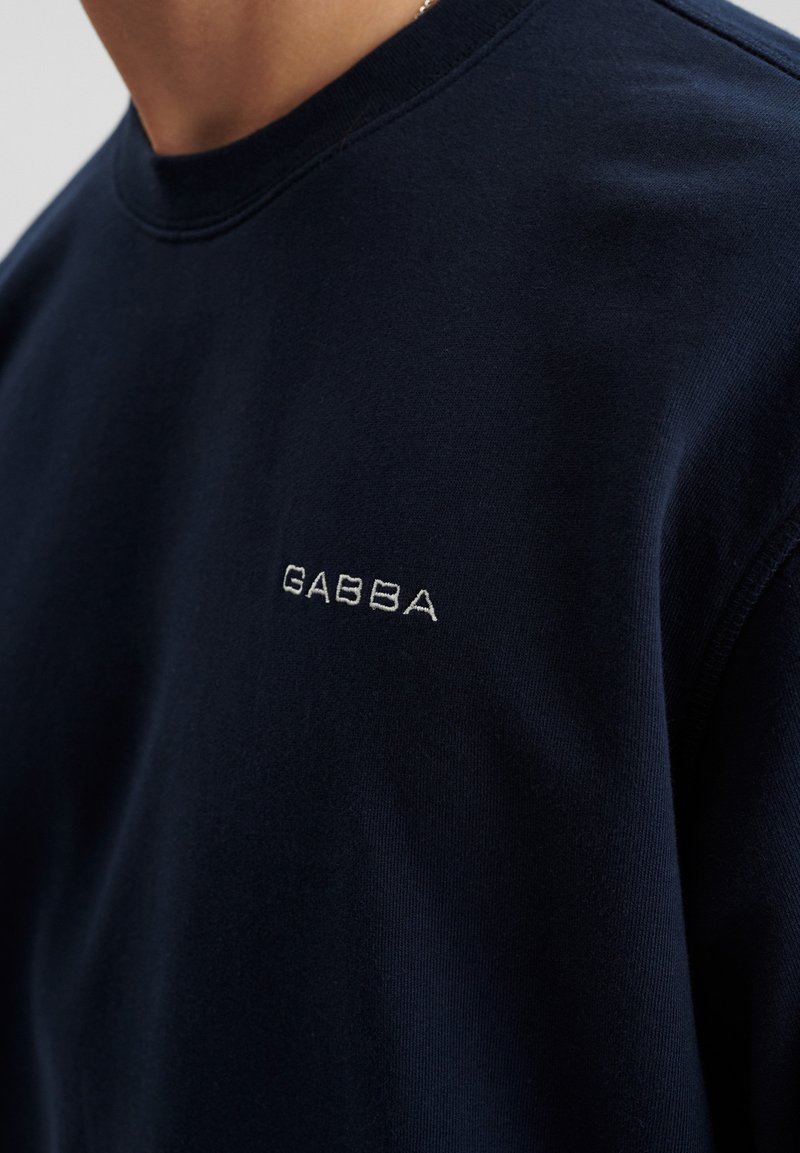 Marineblauwe sweater van zacht materiaal, met een ronde halslijn en een klein wit geborduurd logo op de borst.