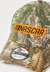 Kamuflázs baseball sapka állítható mérettel. Hímzett narancs 'NASCAR' logóval és zöldes, barnás színárnyalatú textúrájú anyaggal rendelkezik.