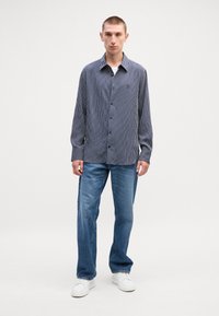 Μακρυμάνικο πουκάμισο ναυτικού με ρίγες, με κουμπιά μπροστά και γιακά, συνδυασμένο με ανοιχτό μπλε τζιν ίσιας γραμμής και λευκά αθλητικά παπούτσια. Απλή φόντο.