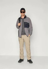 Herre grå fleecejakke med marineblå lomme, kombinert med grå t-skjorte og beige cargobukser, iført svarte sko og solbriller.