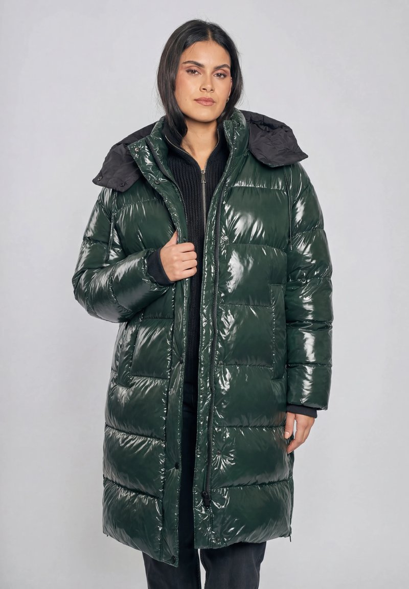 Kobieta ubrana w długi, błyszczący ciemnozielony puchowy płaszcz z czarnym kołnierzem, trzymająca przedni zamek błyskawiczny, na szarym tle.