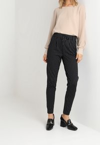 Hellbeige Bluse mit langen Ärmeln, kombiniert mit schwarzen, gestreiften, maßgeschneiderten Hosen. Schwarze Pumps verleihen dem Outfit einen eleganten Touch.