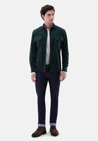 Green corduroy shirt with two chest pockets, worn over a white T-shirt, paired with dark denim jeans and brown lace-up shoes.