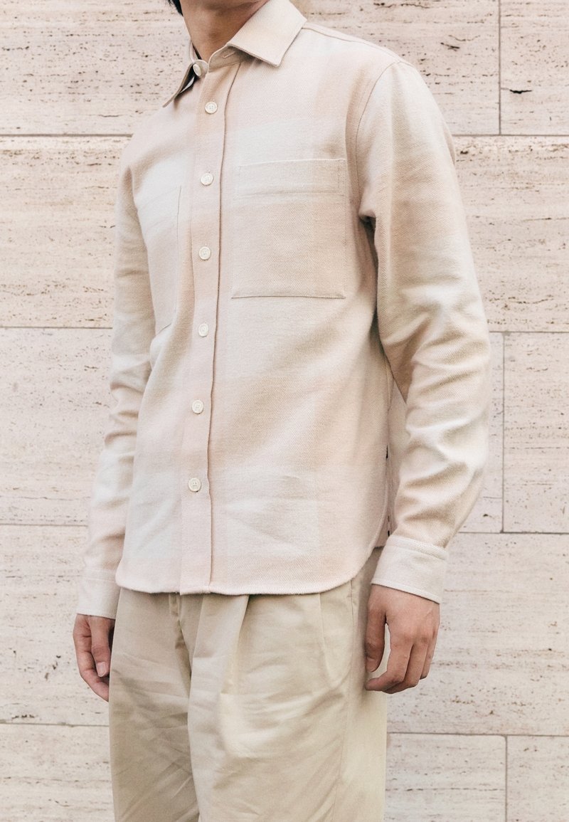Homme portant une chemise beige à manches longues boutonnée avec poches avant et un pantalon plissé de couleur claire, debout contre un mur carrelé.