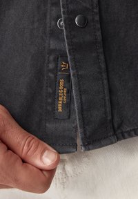 Giacca in denim nera con bottoni a pressione, finitura testurizzata e un'etichetta con "DURABLE GOODS" in oro sulla cucitura interna.