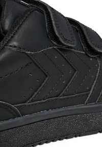 Svart idrottssko med överdel i texturerad läder, kardborreband och gummisula med synligt mönster. Märkt med perforeringar och zigzag-design.
