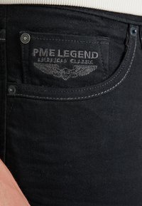 Crne traperice od denima s istaknutom PME Legend etiketom, teksturiranim šavovima i malim metalnim dugmetom na prednjoj džepu.