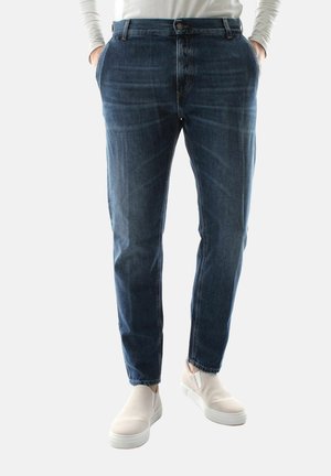 Pantaloni in denim blu scuro con vestibilità dritta, tasche frontali e chiusura con bottoni. Indossati con scarpe slip-on di colore chiaro.