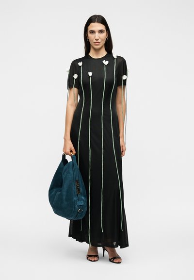 Femme portant une robe maxi noire avec des appliques de fleurs blanches et des lignes verticales vertes, tenant un grand sac à main bleu en daim.