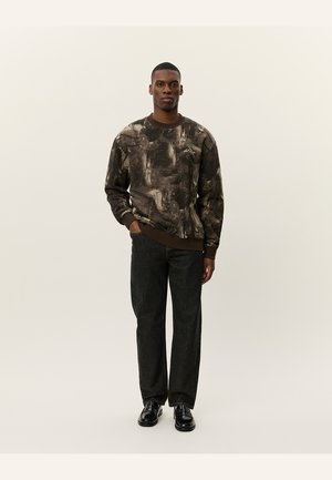 Sudadera para hombre en marrón oscuro con patrones abstractos en colores claros; cuello redondo, puños de canalé, combinada con pantalones de pierna ancha negros y zapatos.
