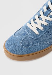 Tenisówki z denimu z jasnoniebieską cholewką, białymi sznurowadłami i brązową gumową podeszwą. Charakteryzują się przeszyciami i zaokrąglonym kształtem noska.