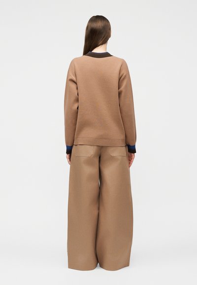 Femme aux longs cheveux bruns vue de dos, portant un pull marron avec des poignets bordés de bleu et un pantalon large couleur beige.