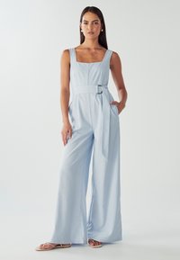 Ženska z dolgimi temnimi lasmi, oblečena v svetlo modro brezrokavno kombinezono z pasom, širokimi nogami hlač in ravnimi sandali, stoji pred belo ozadjem.