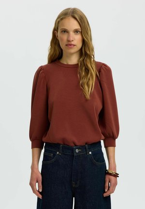Woman with long wavy blonde hair wearing a rust-colored puff sleeve top and dark blue jeans, standing against a plain light background.