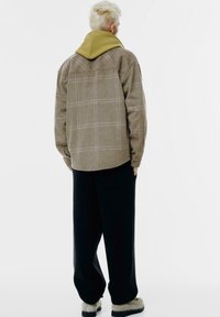 Ruitjen bruin wollen shirtjasje met een mosterdkleurige capuchon, losse zwarte broek en lichtkleurige schoenen. Minimalistisch ontwerp, neutrale tinten.