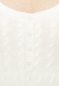 Λευκή πλεκτή ζακέτα με σχέδιο καλώδιου πλέξης, στρογγυλή λαιμόκοψη και πέντε κουμπιά που μοιάζουν με μαργαριτάρια στη μέση μπροστά.