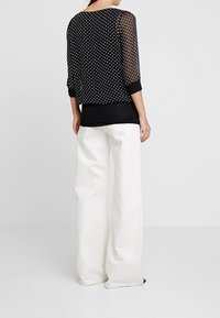 Schwarze transparente Bluse mit weißen Polka-Dots, dreiviertellange Ärmel, kombiniert mit lockeren weißen Hosen. Minimalistisches Design, ohne Verzierungen.