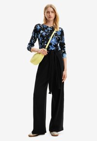 Pantaloni neri lunghi con vita legata, abbinati a una maglietta a maniche lunghe nera e blu con motivi floreali e una borsa crossbody gialla. Scarpe flat metalliche.