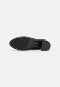 Chaussure en cuir noir avec un bout arrondi et un talon carré. La semelle présente un motif texturé pour une meilleure adhérence. Logo visible sur la semelle.