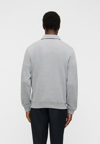 Homme portant un pull gris clair à manches longues avec col, vu de dos, associé à un pantalon noir.