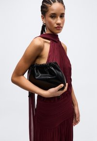 Ung kvinde med fletninger iført en ærmeløs bordeauxrød kjole, der holder en sort lædertaske med snøre mod en ensfarvet baggrund.