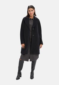 Lange wolblend jas in zwart met een gekleed kraag, twee voorzakken en een relaxte pasvorm, over een grijze button-up jurk en kniehoge zwarte laarzen.