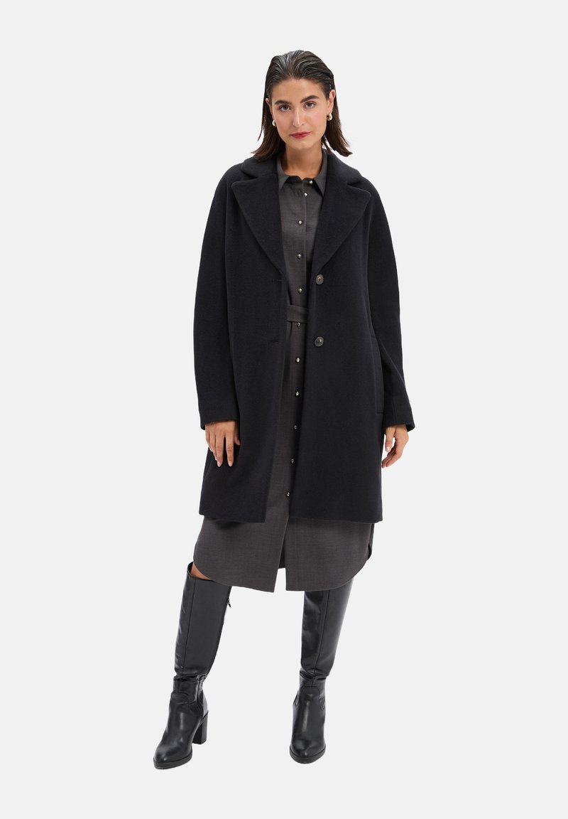 Lange wolblend jas in zwart met een gekleed kraag, twee voorzakken en een relaxte pasvorm, over een grijze button-up jurk en kniehoge zwarte laarzen.