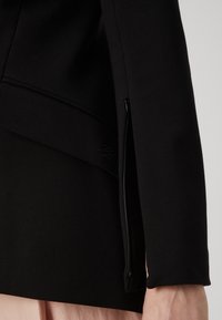 Chaqueta estructurada negra con un ajuste entallado, bolsillo lateral con cremallera y textura suave con un sutil detalle de logo bordado.