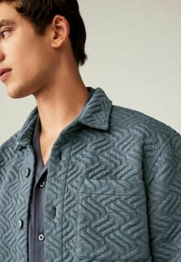 Gesteppte Jacke in Schiefblau mit einem strukturierten geometrischen Muster, ausgestattet mit einem spitzen Kragen und einer Knopfleiste.