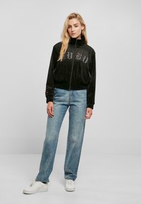 Chaqueta de terciopelo negro con cremallera y puños elásticos, con logo impreso en el pecho; llevada con vaqueros rectos de color azul claro y zapatillas blancas.
