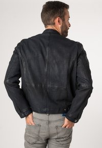 Giacca in pelle nera con finitura texturizzata, dotata di colletto rialzato, maniche aderenti e dettagli con bottoni a pressione. Indossata con pantaloni in denim grigi.