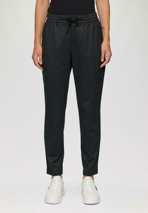 Osoba stojící na světlém jednobarevném pozadí, oblečená v černých a šedých károvaných kalhotách se stahovací šňůrkou a bílých teniskách.