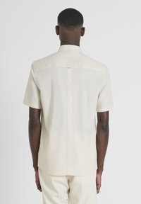Camisa de lino beige de manga corta con un diseño de espalda recta, que presenta una sutil tapeta de botones y líneas limpias y minimalistas.