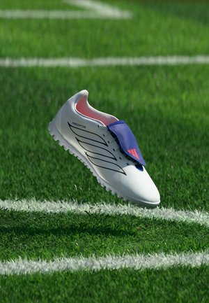 Scarpa da calcio bianca con linguetta blu e design nero che fluttua sopra il campo in erba artificiale verde con linee bianche del campo.