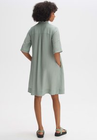 Hellgrünes kurzärmeliges Kleid mit gerippter Textur, knielang, mit seitlichen Taschen und abgerundetem Saum, von hinten gezeigt.