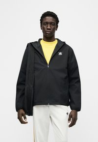 Νεαρός άνδρας όρθιος, φοράει μαύρο μπουφάν Adidas με φερμουάρ, κίτρινη μπλούζα, λευκό παντελόνι Adidas και κρατάει μαύρη τσάντα ώμου.