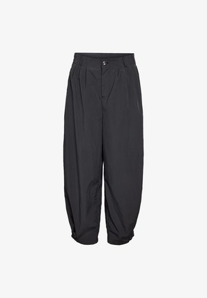 Kvinde står iført en hvid ærmeløs crop top, løse sorte bukser med høj talje og sandaler med hæl mod en ensfarvet lys baggrund.