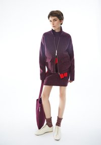 Burgunderfarbener Strickjacke mit Reißverschluss und zwei vorderen Taschen über einem Rock mit roten Akzenten. Weiße Schuhe und passende Tragetasche, strukturierte Socken.